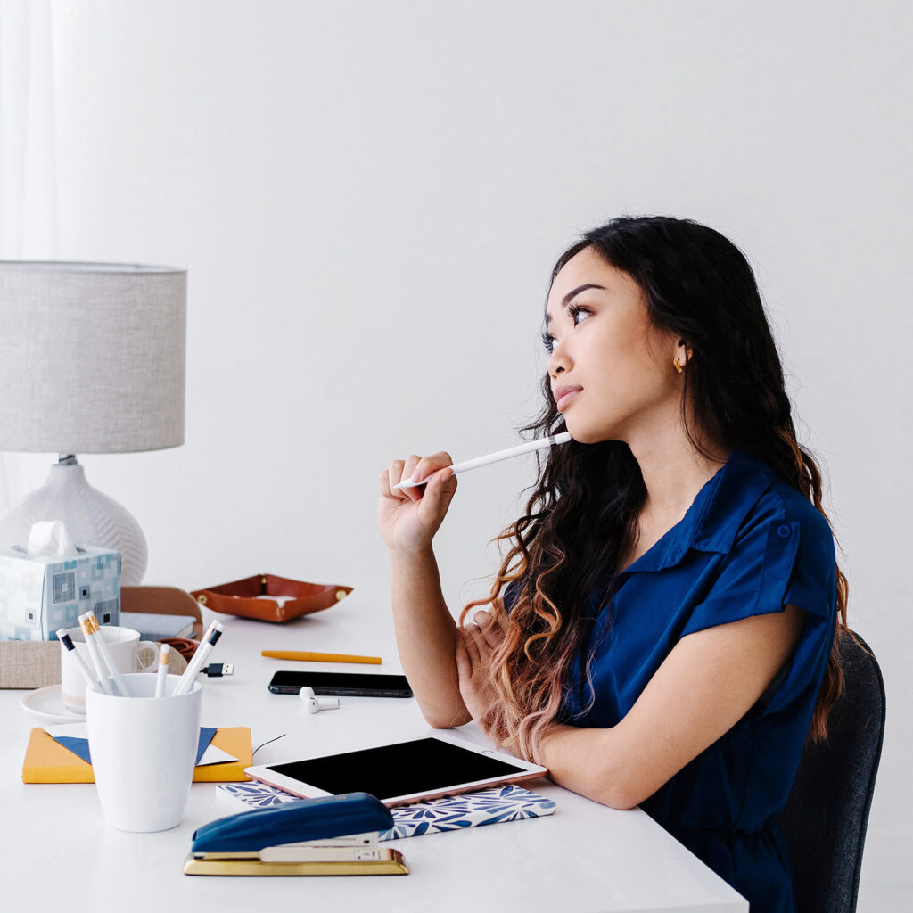 Woman sitting at desk thinking with pencil on chin.